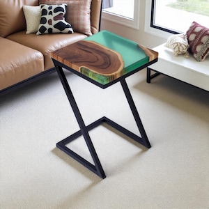 May include: A modern side table with a unique design. The tabletop features a combination of natural wood and a clear, teal-colored resin. The table has a black metal Z-shaped frame. This accent table is a stylish addition to any living room.