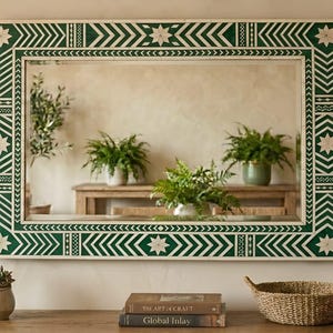 Könnte beinhalten: Rechteckiger Spiegel mit einem grün-weißen geometrischen Rahmen. Der Rahmen weist Zickzack- und Sternmuster auf. Der Spiegel reflektiert Pflanzen und einen Holztisch. Eine kleine Sukkulente im Topf, Bücher und ein geflochtener Korb stehen auf dem Tisch.