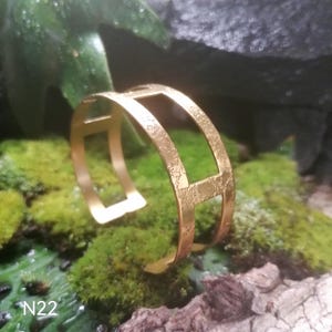 以下が含まれることがあります： テクスチャ加工された表面とオープンデザインのゴールドトーンのカフブレスレット。 ブレスレットは長方形の切り抜きがあり、緑色の苔の上に表示されています。 テキスト「N22」が左下に表示されています。