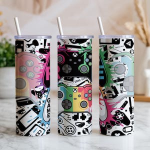Puede incluir: Tres vasos cilíndricos con tapas transparentes y pajitas blancas, con un estampado gráfico vibrante de varios mandos de videojuegos, salpicaduras de pintura de colores y el texto "GAME OVER" en letras negras sobre un fondo blanco.