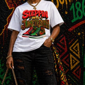 Op de afbeelding: Wit T-shirt met de tekst "Steppin' Into Juneteenth" in rood, groen en geel. Het shirt heeft een afbeelding van rode en groene sneakers. De persoon draagt een zwarte ripped jeans en een gouden ketting.