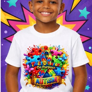 May include: A white t-shirt featuring a colorful graphic design with puzzle pieces, butterflies, and the text "Built Different On Purpose" and "Autism". The design is set against a purple background with star and lightning bolt accents.