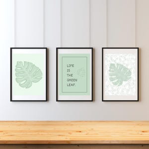 Può includere: Tre stampe artistiche incorniciate con un tema di foglia di monstera. Le stampe sono in cornici nere con un bordo bianco. Una stampa mostra un'illustrazione di foglia, un'altra ha il testo "LIFE IS THE GREEN LEAF", e la terza ha un motivo a foglia.