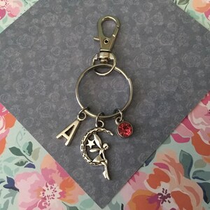 May include: Silver keychain with a lobster clasp, a large ring, and three charms. Charms include the letter "A", a fairy on a crescent moon, and a small pink gem. The keychain is on a grey floral background.