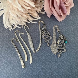 May include: A collection of five silver-toned metal bookmarks. Three are simple curved rods with textured patterns or a feather shape. Two feature dangling charms including snakes, flowers, a letter B, a letter N, and a small blue crystal bead.