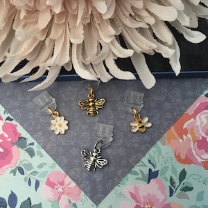 May include: Four decorative earring charms with bee and flower designs. The charms are gold and silver-toned metal with white, yellow, and black enamel details. The charms are attached to clear earring clips.
