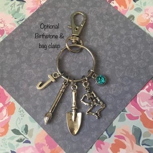 May include: Silver-coloured keyring with a lobster clasp, a large ring, and several charms. Charms include a letter "J", a paintbrush, a trowel, a skeleton, and a teal-coloured gemstone. The text "Optional Birthstone & bag clasp" is visible.