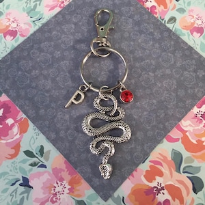 May include: Silver-coloured keychain with a detailed snake charm, a red gemstone, and a letter "P" pendant. The keychain includes a clasp and a key ring. The background is a floral pattern.