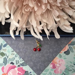 May include: A gold cherry charm with red enamel cherries and a green leaf. The charm hangs over a blue and floral patterned background, with a large, light pink flower in the upper portion of the image.