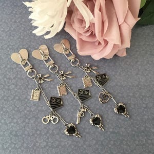 May include: Silver keychains with heart-shaped clasps and various charms, including books, playing cards, handcuffs, and a gun. The charms are connected by delicate chains. The background features pink and white flowers.