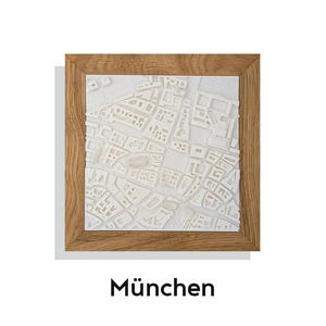 May include: A framed, three-dimensional white map of a city, set against a white background. The map is encased in a natural wood frame. The word "München" is printed in black at the bottom of the frame.