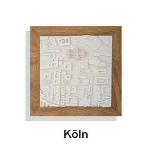 May include: A framed, three-dimensional white architectural model of a city, possibly Cologne, Germany. The model is set within a natural wood frame. The word "Köln" is printed in black at the bottom of the image.