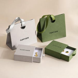 Può includere: Due sacchetti regalo e due scatole per gioielli. I sacchetti sono grigio chiaro e verde oliva con manici in nastro abbinati. Le scatole sono grigio chiaro e verde oliva, con il nome del marchio "PUPPYSEW" stampato sopra. Le scatole sono aperte, rivelando gioielli dorati e verdi.