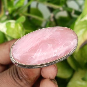 May include: A silver ring featuring a large, oval-shaped pink gemstone. The gemstone has a swirling, marbled appearance. The ring is held against a backdrop of green foliage.
