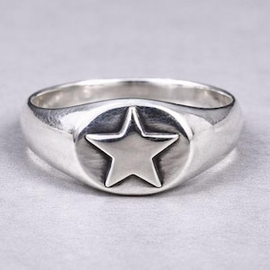 Puede incluir: Anillo de plata con diseño de estrella. El anillo tiene una banda redondeada y una cara circular con una estrella en relieve en el centro. El anillo es de metal brillante.