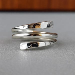 May include: A silver-colored wrap ring with a spiral design. The ring has a smooth, polished finish with a hammered texture on the ends. The ring is shown on a gray surface with a blurred brown background.