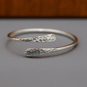 Puede incluir: Una pulsera de puño plateada con textura martillada en los extremos. La pulsera tiene un diseño abierto, con los extremos ligeramente superpuestos. El metal tiene un acabado brillante. La pulsera se muestra sobre una superficie gris.