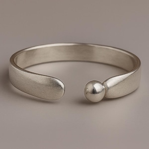 Könnte beinhalten: Ein silberfarbenes Armreif-Armband mit offenem Design. Das Armband hat ein abgerundetes, offenes Band mit einem kleinen, kugelförmigen Akzent auf einer Seite. Das Metall hat ein mattes Finish, und der Hintergrund ist ein neutrales Grau.