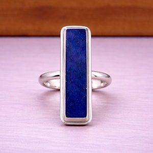 May include: A silver ring featuring a rectangular, deep blue stone. The stone is set in a silver bezel, and the ring has a simple, elegant design. The ring is displayed on a light purple surface.