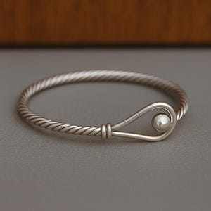 Puede incluir: Un brazalete plateado con un diseño de cuerda trenzada. El brazalete presenta un cierre de gancho y ojo con una pequeña bola redonda. El brazalete está sobre una superficie gris.
