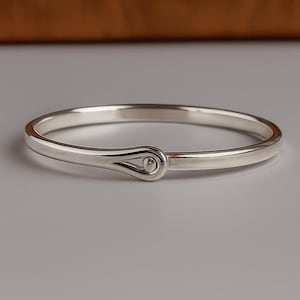 Puede incluir: Un brazalete plateado con cierre de gancho y ojo. El brazalete tiene un acabado liso y pulido y un diseño simple y elegante. El cierre de gancho presenta una forma de lágrima con un pequeño detalle redondo.