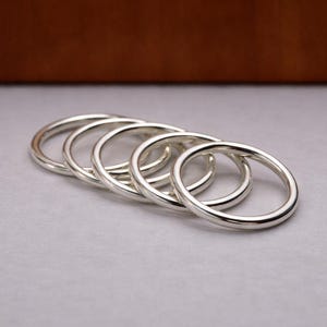 May include: Five silver rings are displayed on a grey surface. The rings are circular and have a smooth, polished finish. The background is a blurred brown colour.