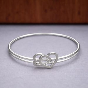 Op de afbeelding: Een zilverkleurige armband met twee met elkaar verbonden hartvormige bedels. Eén hart heeft een bliksemontwerp. De armband ligt op een grijze ondergrond.