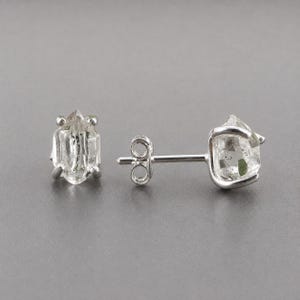 Peut inclure: Une paire de boucles d'oreilles à clous avec des cristaux de quartz bruts, transparents et hexagonaux, sertis dans des griffes en métal argenté. L'une est vue de face, l'autre de profil, montrant la tige et le fermoir papillon.