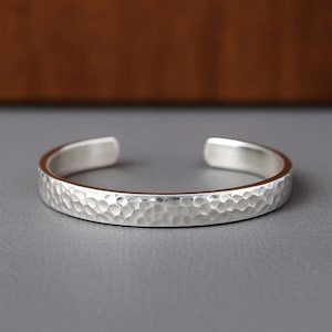 Pode incluir: Uma pulseira de punho prateada com textura martelada. O design aberto apresenta uma superfície texturizada e um interior polido. Esta joia é um acessório simples, mas elegante.