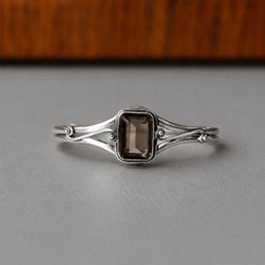 May include: A silver ring featuring a rectangular, emerald-cut smoky quartz gemstone set in the center. The band is crafted from polished silver with a split-shank design that curves outward, accented by small silver spheres on each side of the stone.