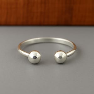 Puede incluir: Una pulsera de puño abierta de color plateado con una banda lisa y redondeada. Cada extremo de la pulsera presenta una pequeña esfera pulida. La pulsera se muestra sobre una superficie gris, con un fondo marrón.