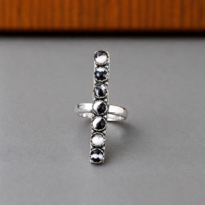Può includere: Un anello in argento con una barra verticale incastonata con sette pietre rotonde dal motivo bianco e nero. Le pietre sono disposte in un'unica colonna, ciascuna tenuta in una montatura a festone, su una fascia in argento liscia.
