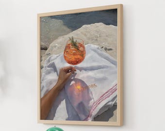 Könnte beinhalten: Gerahmter Kunstdruck eines Cocktails auf einem weißen Handtuch. Das orangefarbene Getränk befindet sich in einem großen Glas, garniert mit Rosmarin. Das Handtuch hat einen roten Streifen und Text. Im Hintergrund sind Felsen und Wasser zu sehen.
