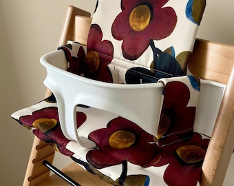 Peut inclure: Une chaise haute en bois équipée d'une barre de sécurité en plastique blanc et d'un coussin rembourré. Le coussin présente un motif floral à grande échelle dans des tons rouge foncé, bleu marine et or sur un fond de couleur crème.