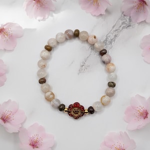 May include: A beaded bracelet featuring smooth, round, translucent off-white and marbled beige beads interspersed with smaller, dark brown textured spacer beads. A single, central, deep red flower-shaped bead with gold-tone accents sits at the bottom.