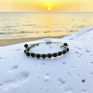 May include: A handmade bracelet featuring dark blue beads strung on a light green cord. The bracelet is displayed on a white, textured surface with a beach and sunset in the background. The beads are evenly spaced.