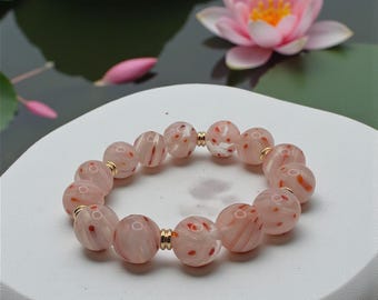 Pode incluir: Uma pulseira de contas rosa com salpicos vermelhos e brancos e espaçadores dourados. A pulseira está sobre uma superfície branca, com um nenúfar rosa e folhas de nenúfar ao fundo.