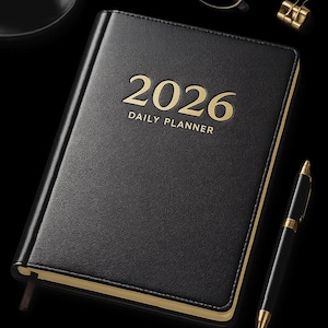 May include: A black 2026 daily planner with gold lettering and trim, a black pen, gold-rimmed glasses, and a gold paper clip. A black coffee cup and saucer are also present. The items are arranged on a black surface.