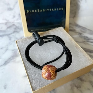 May include: A brown wooden bead with a floral design is on a black elastic bracelet. The bracelet is in a brown box with the text "BlueSagittarius" on the lid.