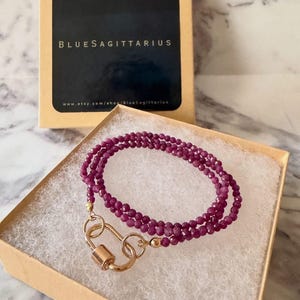 May include: A multi-strand bracelet made of small, faceted, deep magenta-colored beads. The bracelet features a gold-toned metal carabiner-style clasp. It is presented in a tan cardboard box with a black label reading BLUESAGITTARIUS.