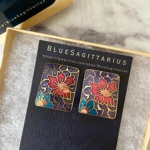May include: A pair of rectangular stud earrings with a floral design. The earrings feature a red flower with blue and pink accents on a dark purple background, framed by a gold border. The text "BlueSagittarius" is visible.
