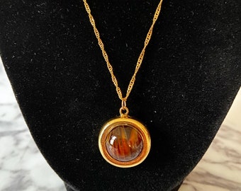 May include: A gold-colored necklace with a round pendant. The pendant has a gold frame and a brown, amber-like center. The chain is a twisted rope design. The necklace is displayed on a black velvet surface.