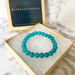 May include: A teal blue beaded bracelet in a small, brown gift box. The box has a label that reads "BlueSagittarius" and a website address.
