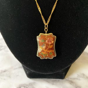 May include: A gold-toned necklace with a rectangular pendant featuring a floral and bird design. The pendant has a reddish-brown background with a gold border. The chain is a delicate, twisted rope style.