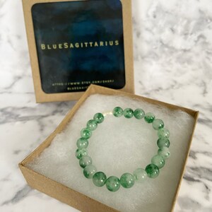 K&ouml;nnte beinhalten: Ein gr&uuml;nes Jade-Armband mit einem silbernen Verschluss in einer braunen Schachtel. Die Schachtel tr&auml;gt ein Etikett mit der Aufschrift "BlueSagittarius".