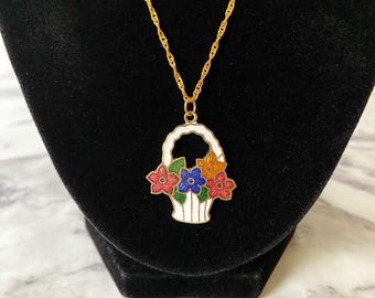 May include: A gold-colored necklace with a pendant shaped like a white basket filled with colorful flowers. The flowers are red, blue, and orange. The necklace is displayed on a black velvet background.