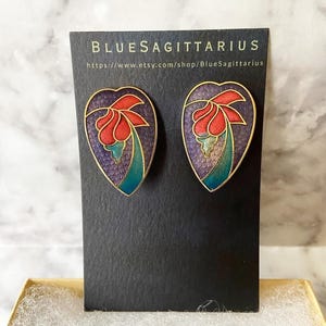 May include: A pair of heart-shaped earrings with a floral design. The earrings are made of gold metal and have a purple, blue, and red enamel finish. The earrings are on a black card with the text "BLUESAGITTARIUS" and a website address.