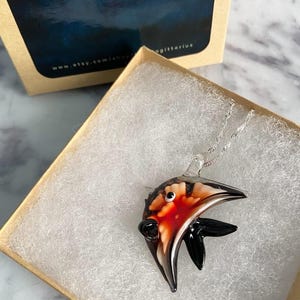 May include: A glass pendant necklace in a gift box. The pendant is shaped like a fish with a red, black, and white design. The necklace has a silver chain. The gift box is tan with the text "BLUESAGITTARIUS" and a website address.