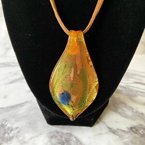 May include: A teardrop-shaped glass pendant necklace with a gold, green, and blue color scheme. The pendant has a textured surface and is suspended from a brown suede cord.