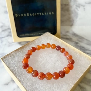 May include: A carnelian bead bracelet displayed in a small, open box with white padding. The bracelet features round, orange and red beads. The box is inside a larger box with the text "BLUESAGITTARIUS" on the front.